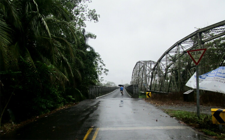 An asphalt road leading over a bridge with forest to one side and arched metal constructions of an older bridge to the other side