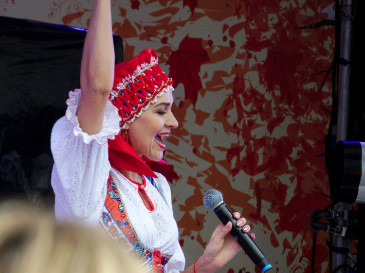 A woman in traditional Russian clothing singing enthusiastically, one arm raised into the air, the other holding a microphone