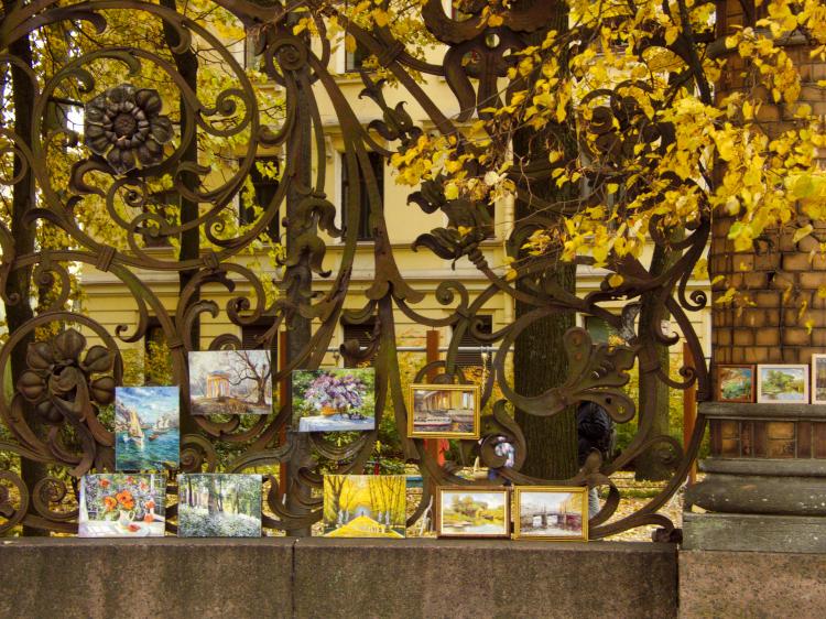 Various small paintings being displayed for sale on an intricate, floral cast-iron fence, with the yellow leaves of autumn trees in the background