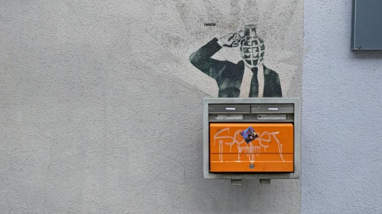 An orange mailbox mounted on a wall with a stencilled graffiti above showing a businessman whose head is replaced by a grenade