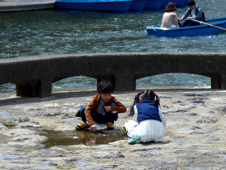 Two kids sitting around a small puddle playing near a lake