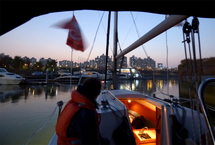 A person in a life jacket sitting on the side of a sailboat on a river running through an evening cityscape
