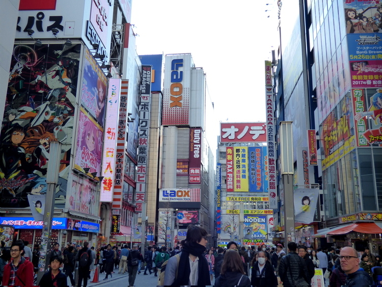 A busy pedestrian area lined with buildings bearing large posters advertising various brands, many with Anime or Manga imagery