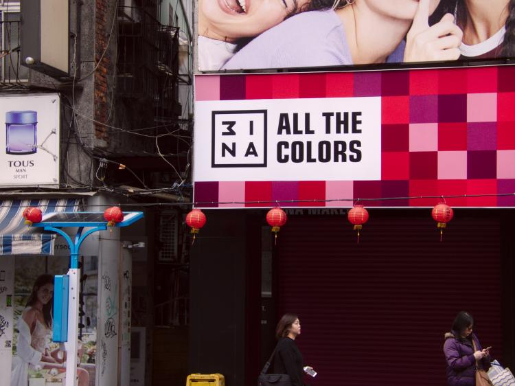 A street-side view with two people walking across and an advertisement poster featuring a red geometric pattern and the text 'all the colors'