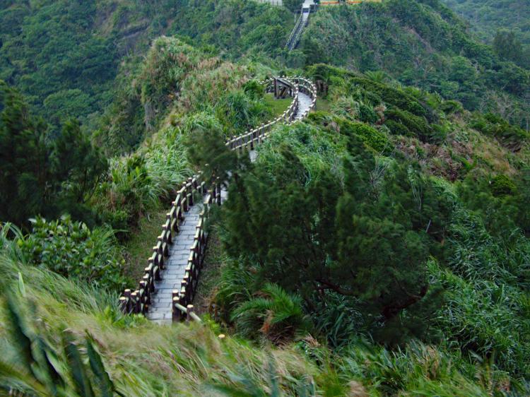 A narrow wooden walkway with railings to either side winding along the ridge of a green hill and out of sight