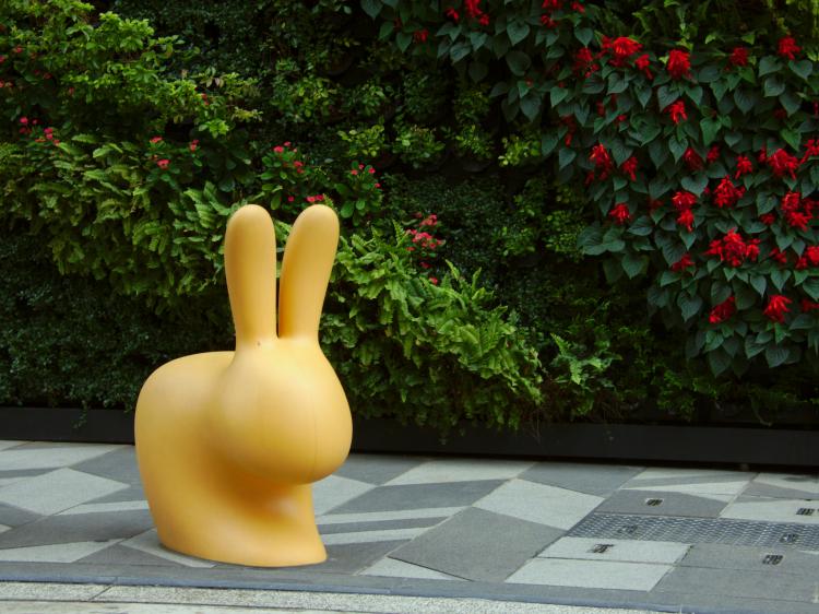 An abstract yellow plastic sculpture of a rabbit on a street in front of an overgrown wall