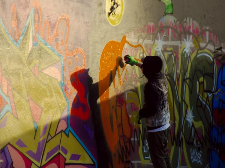 A person in a dark hoodie spraying large orange letters on a wall covered in grafitti