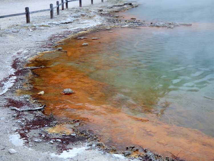 A geothermal pool fading in colour from orange-red to blue with steam rising from it