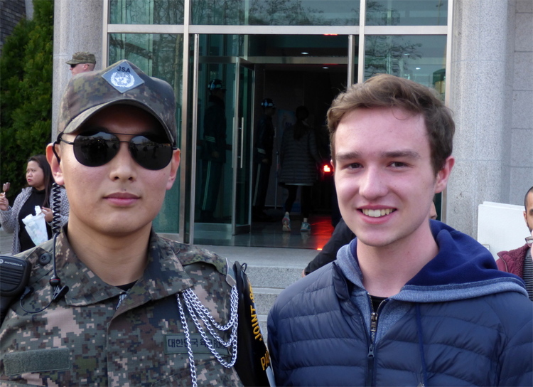 Jan posing for a picture besides a young male South Korean soldier in camouflage uniform and dark sunglasses
