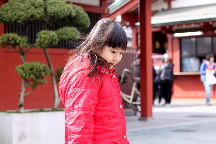 A young girl in a bright red jacket playing on a square in front of some red buildings