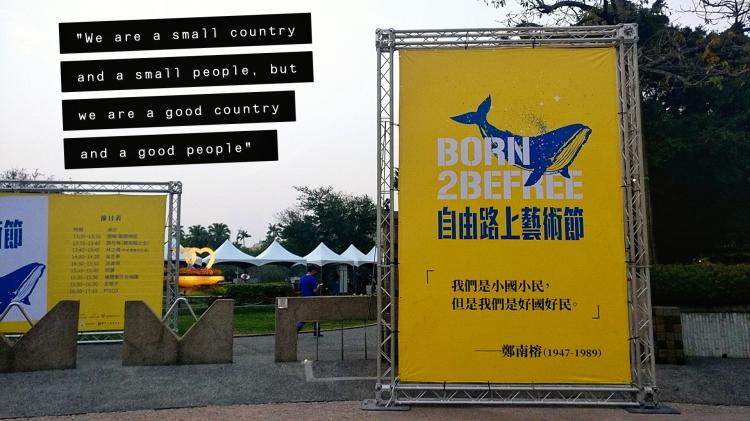 A yellow banner standing in a park bearing the slogan 'Born 2 be free' and a quote in Chinese translating to 'We are a small country and a small people, but we are a good country and a good people'