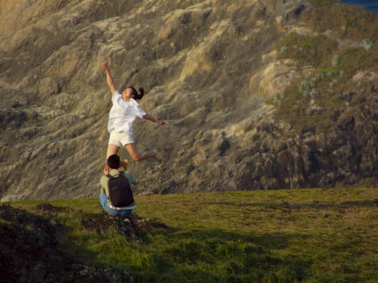A woman jumping into the air with her arms stretched out, hair and clothes blown back by the wind, being photographed by a man kneeling in front of her