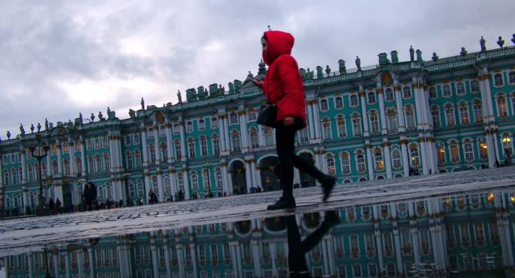 A woman in a red jacked walking across a puddle reflecting a turquoise building