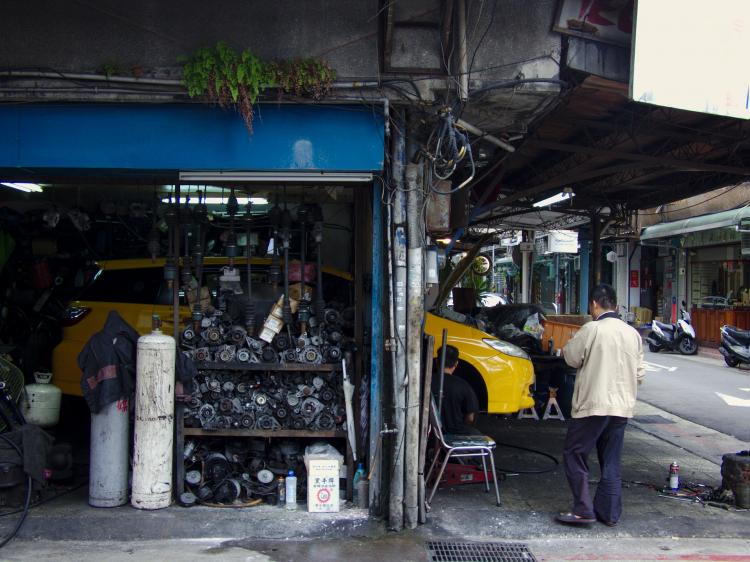 A yellow car parked in an open garage in the ground floor of a building with racks of spare parts all around, with a man looking at the open hood