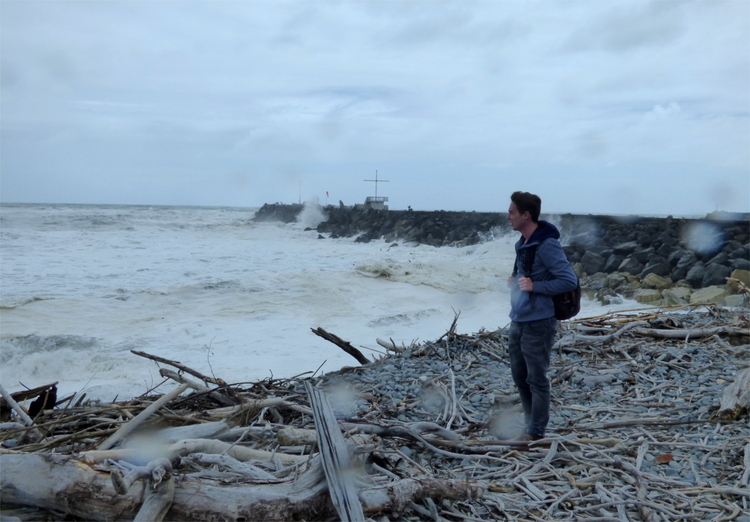 Jan standing on a driftwood-covered shore looking out at the foamy white sea and the grey sky