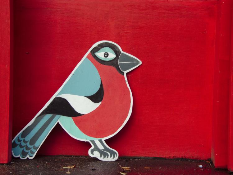 A light blue and red bird painted on a wooden cut-out leaning against a deep red wall