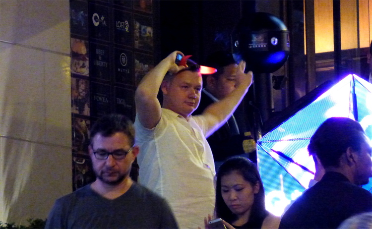 An apparently drunk white man with light-up devil horns on his head standing in a group of people