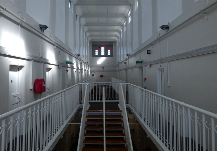 The white-painted interior of an old prison building with a high ceiling and metal railings, cell doors leading off to both sides