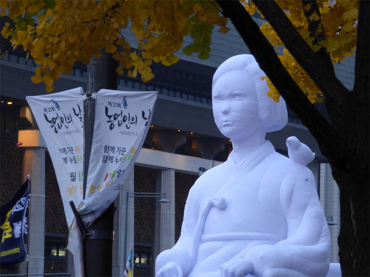 A large white inflatable doll showing a stern-looking woman sitting with a bird on her shoulder under a yellow ginkgo tree