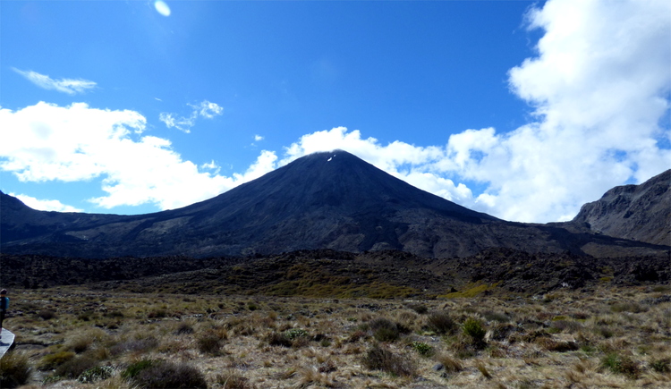 A large, conic black volcano visible in the distance surrounded by white clouds on a blue sky
