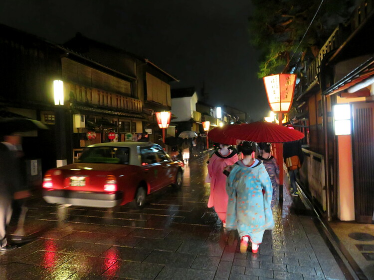 A group of Geisha in brightly coloured Kimonos walking along a rainy street in Kyoto under red umbrellas in the evening, a red-and-white taxi passing them on the left