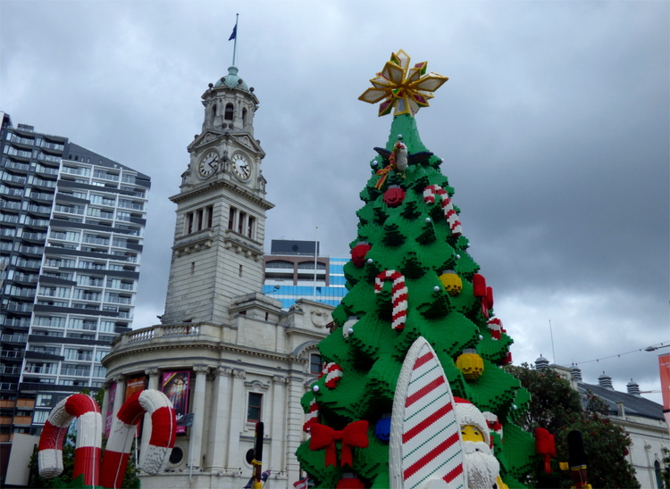 A larger-than-life Lego Christmas tree with matching Lego Santa holding a Surfboard in front of a white stone church tower
