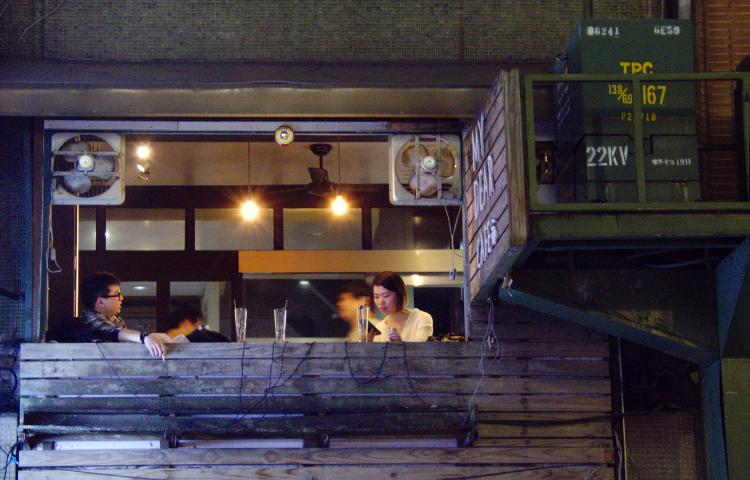 An evening scene showing a man and a woman sitting in a bar on the first floor of a building with its side open to the street, some empty glasses between them