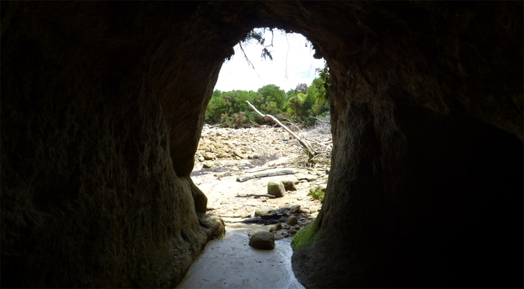 A tall, rounded exit of a person-sized natural tunnel showing the beach outside of it