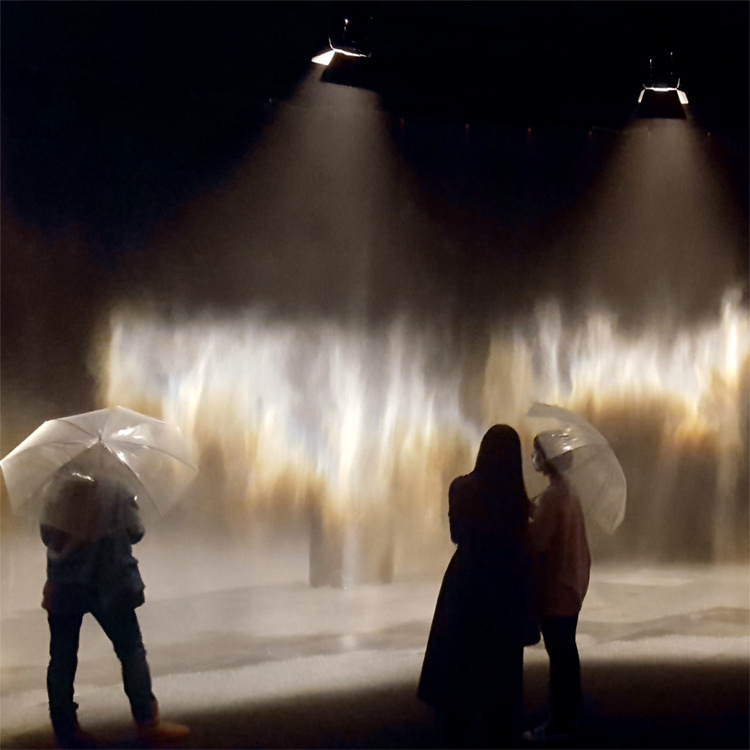 Museum visitors with transparent umbrellas looking at an indoor rainbow created from mist and light