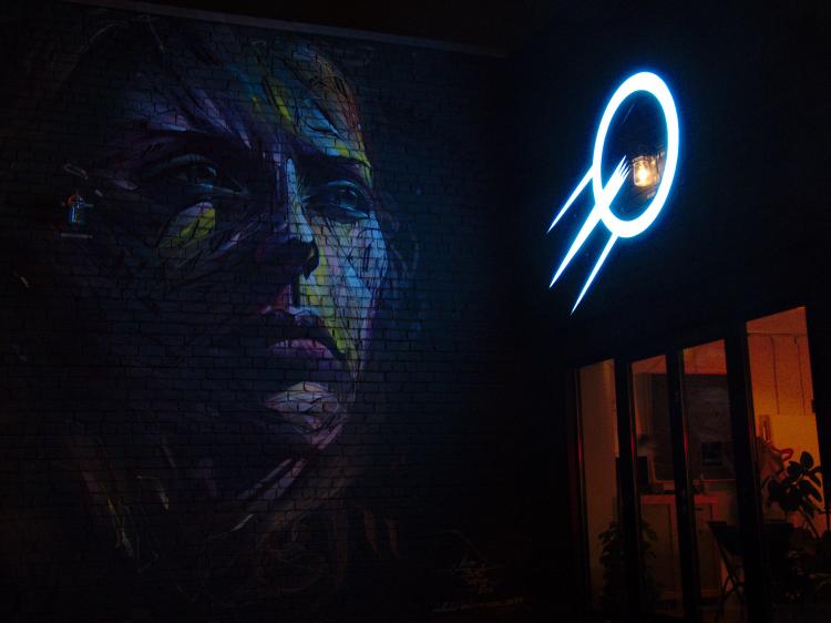 A mural on a brick wall showing a woman's face, photographed at night and illuminated by a nearby neon sign showing a stylized sattelite