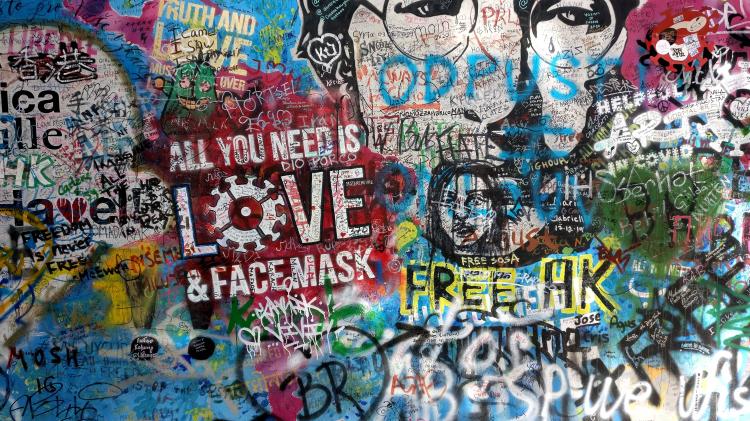 A wall covered in graffiti and tags, featuring a portrait of John Lennon and the slogan 'all you need is love & facemask', with the 'o' in 'love' replaced by a stylized virus