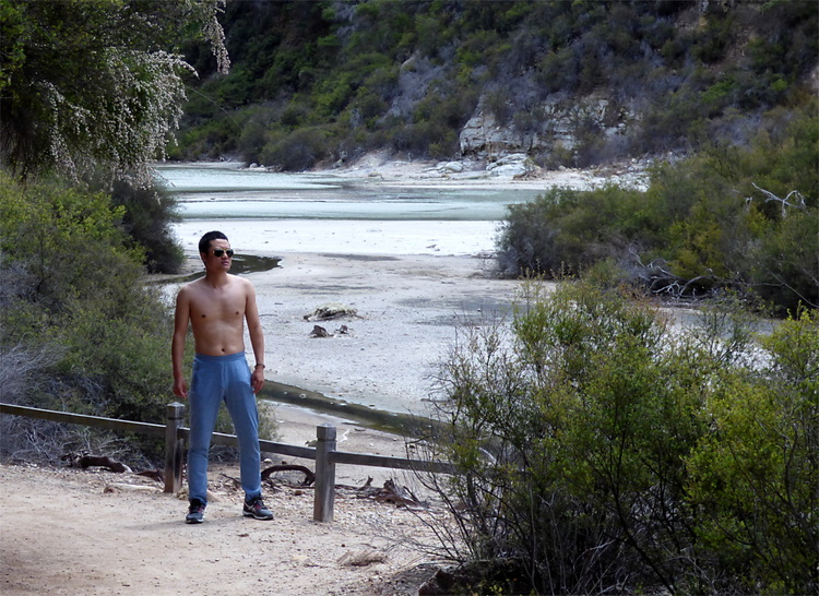 A man with sunglasses poses topless in front of a lake