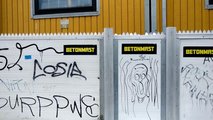 A white temporary construction fence in front of yellow containers. There are many black tags sprayed and drawn on the fence, among them a cartoonish sketch of Munch's "The Scream"