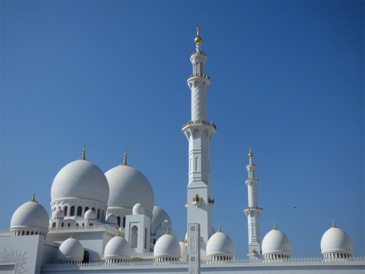 Silhouette of a white stone mosque with many white domes and two minarets against a clear blue sky