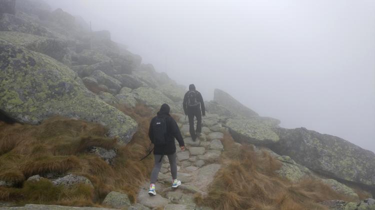 Two people walking a narrow path through a rocky mountain landscape in heavy fog