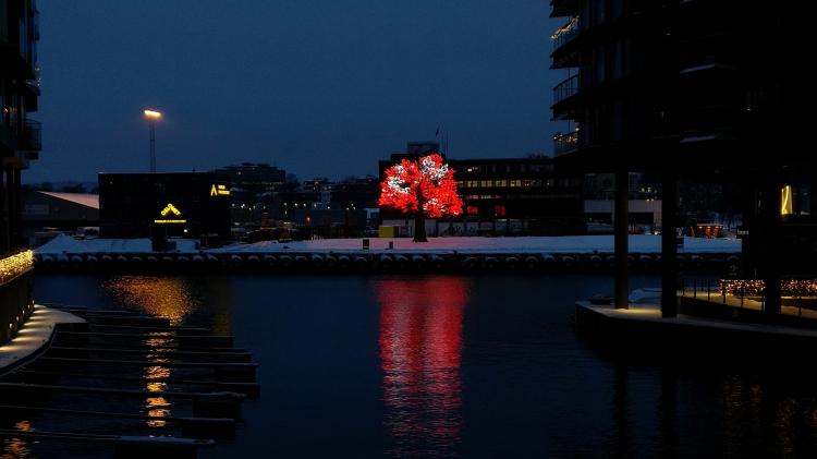 A large, tree-like sculpture made of thousands of lights standing in the snow across the water, its red and white light reflected on it.