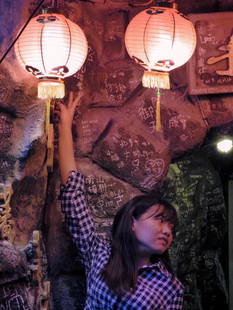 A young woman in a blue plaid shirt posing for pictures underneath some red lanterns, reaching one hand up towards them
