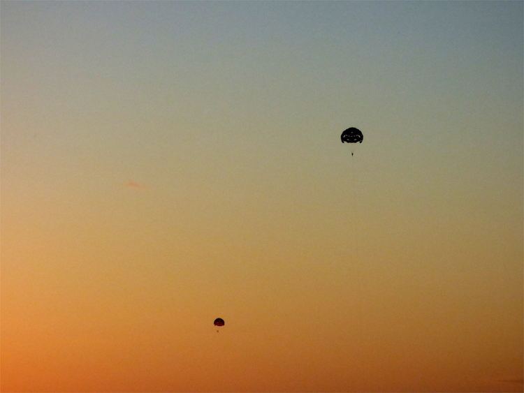Black silhouettes of two parachutes against a sunset sky fading from blue-grey into deep orange