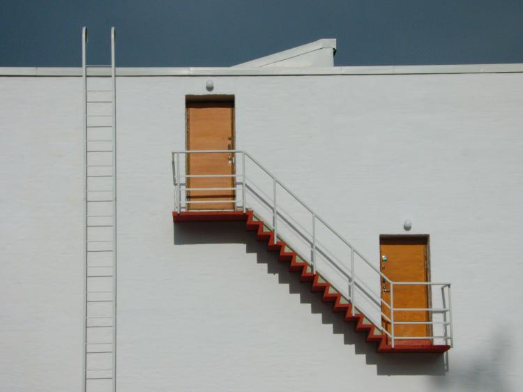 Two orange doors up on the side of a white wall, connected by narrow red stairs, inaccessible by any other means, with a white ladder leading to the roof of the building