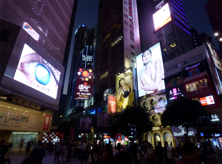 LED advertising screens on a busy public square in the evening, one showing a close-up of a human eye