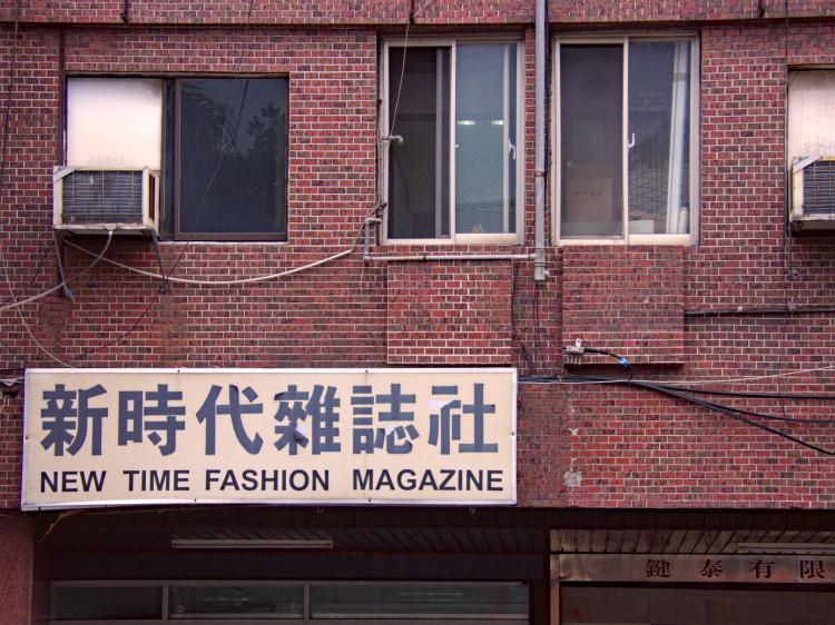 A dark red-brick facade with some windows and AC units, a white rectangular sign with large Chinese writing reading 'New Time Fashion Magazine' underneath