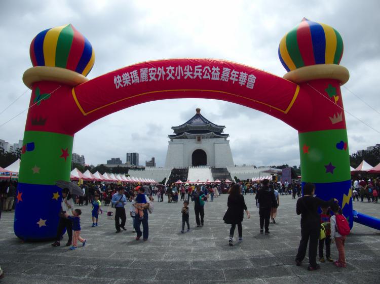 A multi-coloured inflatable gate on a public square hosting some sort of festival, a grand memorial hall visible in the background