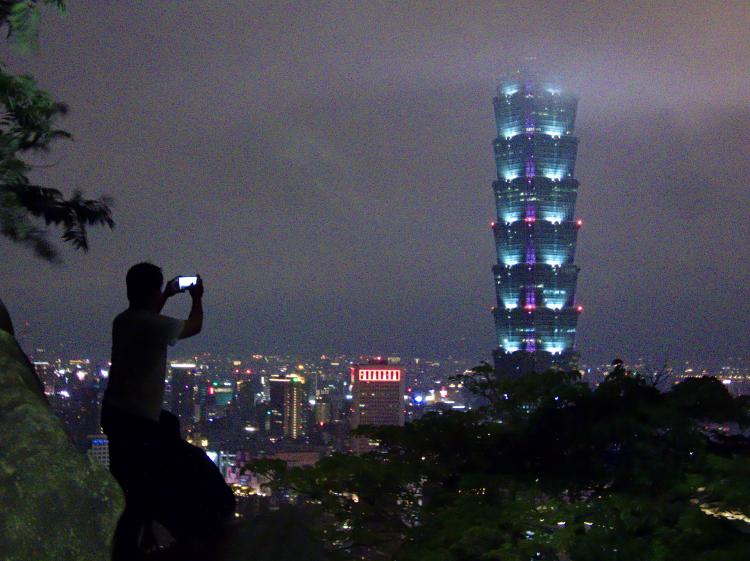 A person standing on a rock taking a picture of the Taipei 101 at night