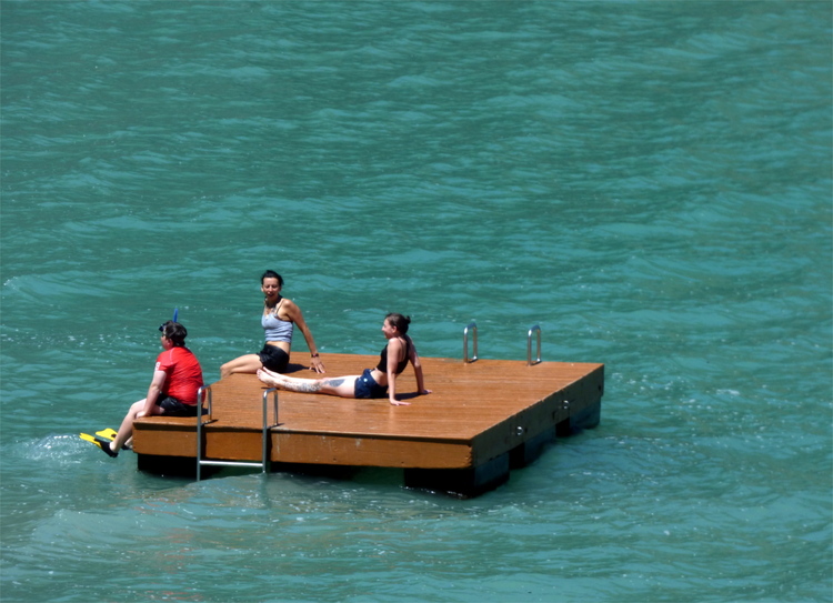 Three people in swimming gear sitting on a square wooden bathing island in turquoise water