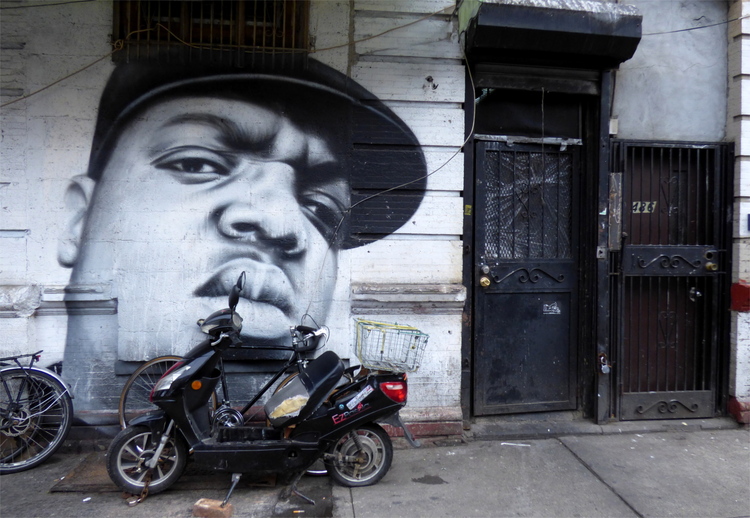 Black-and-white street-art showing the face of a man wearing a cap with a motor scooter parked in front