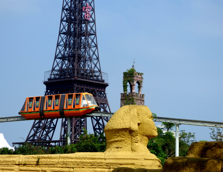 An orange monorail car moving past a replica of the Eiffel tower and the Sphinx of Gizeh with an overgrown church tower in the background