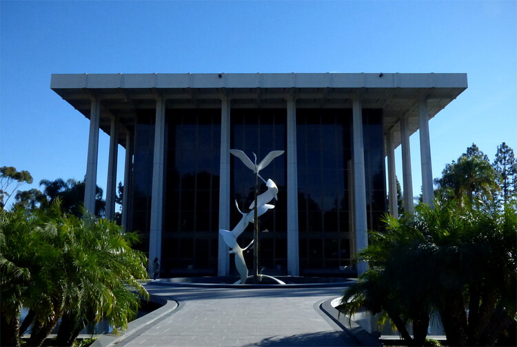 A sculpture of large white birds rising up around a central column in front of a modern building with a slab roof supported by tall columns, almost reminiscent of Greek temples
