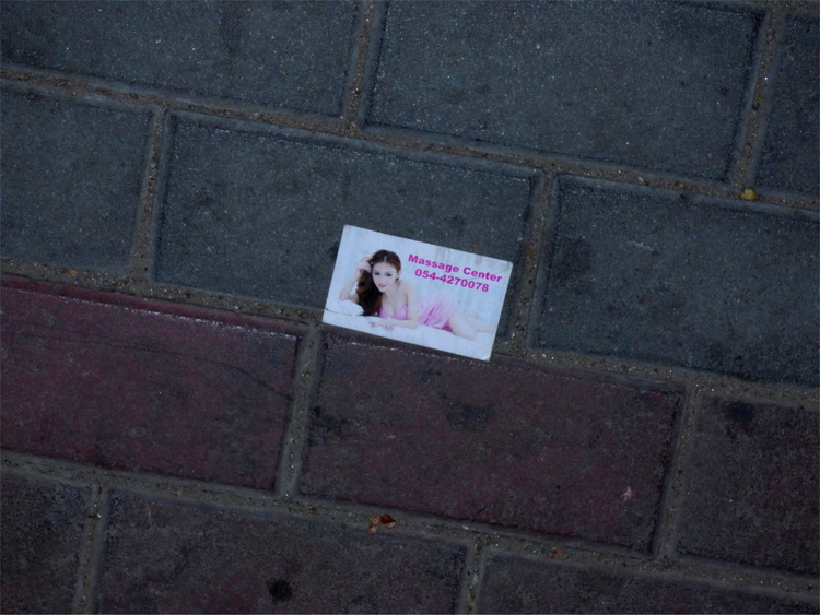 A business card showing a thinly-clothed woman on a bed reading 'Massage Center' with a phone number underneath laying on the street