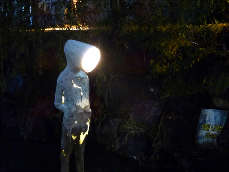 A vaguely humanoid sculpture with light leaking out from inside through cracks in its surface, with a head shaped like a lamp and light emerging from it