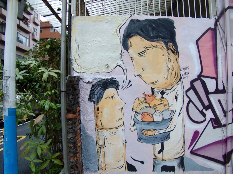 A cartoon-style mural on the side of a wall showing a father and son, the father holding a tub of water balloons, a small signature reading 'Candy Bird'